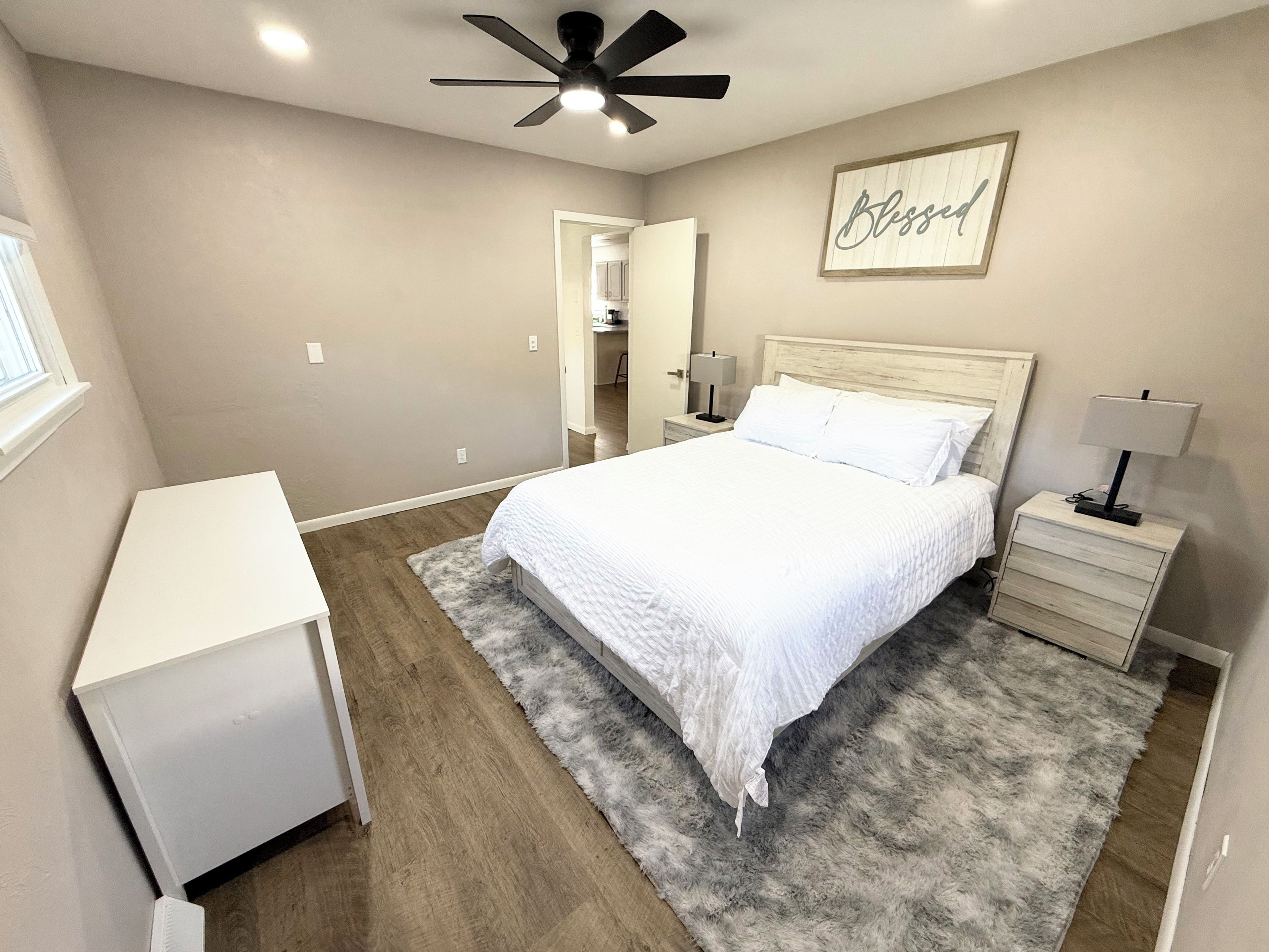 A bedroom with a bed, bedside tables, a desk, a ceiling fan, and a framed sign on the wall.