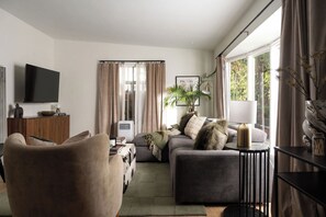 Een moderne woonkamer met een flatscreen televisie, een bank, een fauteuil en een groot raam met gordijnen.