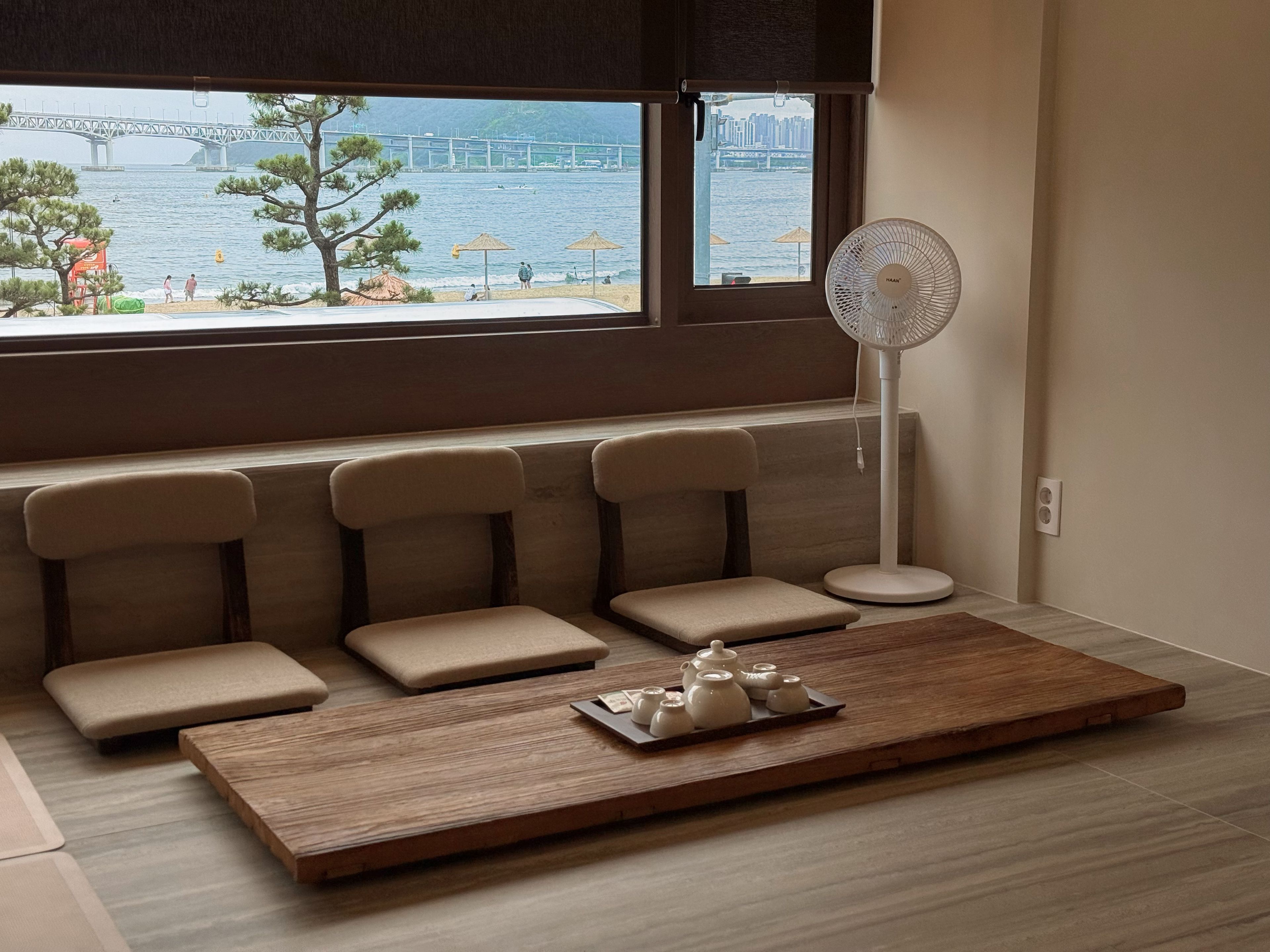 Una habitación con zona de estar junto a una ventana con vistas a un puente y al agua, un ventilador de pie y una bandeja con teteras.