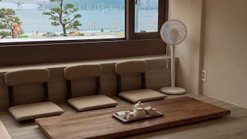 Una habitación con zona de estar junto a una ventana con vistas a un puente y al agua, un ventilador de pie y una bandeja con teteras.