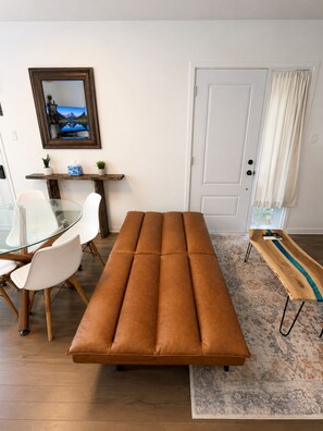 Ein modernes Wohnzimmer mit einem Glastisch, weißen Stühlen, einem braunen Ledersofa, einem Holztisch und einem gemusterten Teppich.