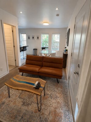 Ein Wohnzimmer mit einer Couch, einem Couchtisch und einem Teppich. Eine Tür ist links, ein Fenster rechts.