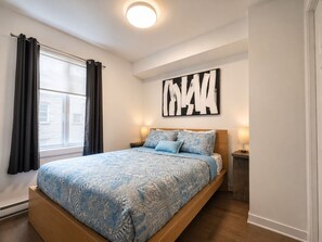 Ein Schlafzimmer mit einem Bett, einem Fenster mit Vorhängen, einem Wandbild und Nachttischlampen.