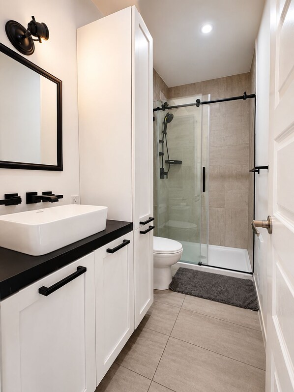 Une salle de bain moderne avec un lavabo blanc, un plan de toilette noir et une douche à l’italienne en verre.
