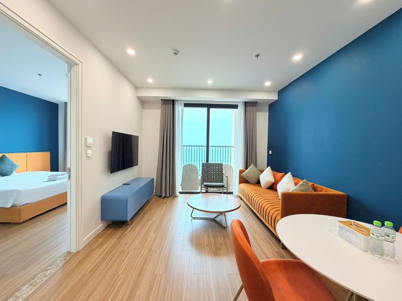 A modern hotel room with a bed, a sofa, a dining table, and a TV.