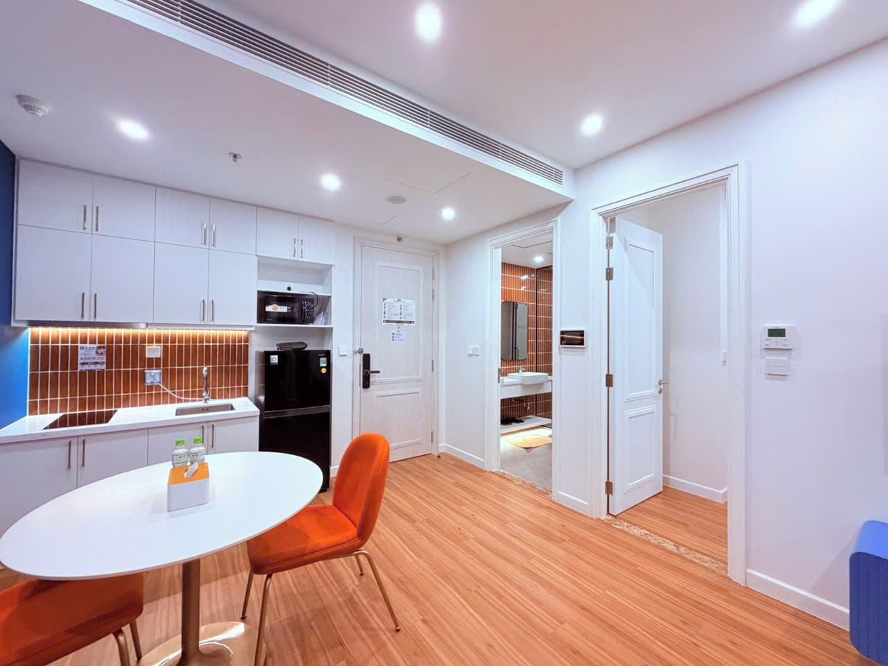 A modern kitchen with white cabinets, a black refrigerator, and a dining area with a white table and orange chairs.