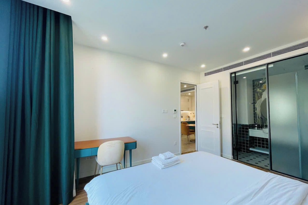 Habitación de hotel con cama, escritorio, silla y baño visible a través de una puerta de cristal.
