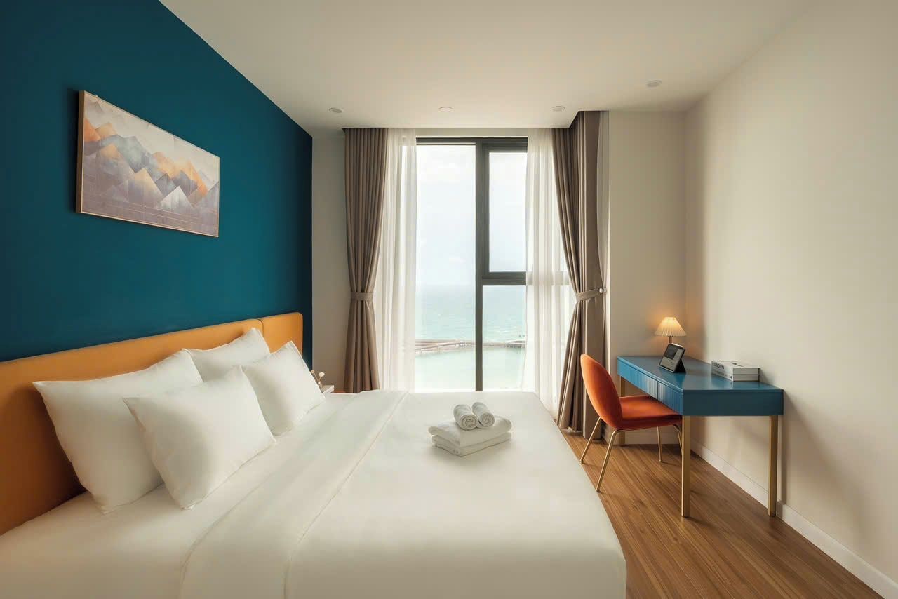 A hotel room with a bed, a desk, a chair, a painting on the wall, and a view of the sea through the window.