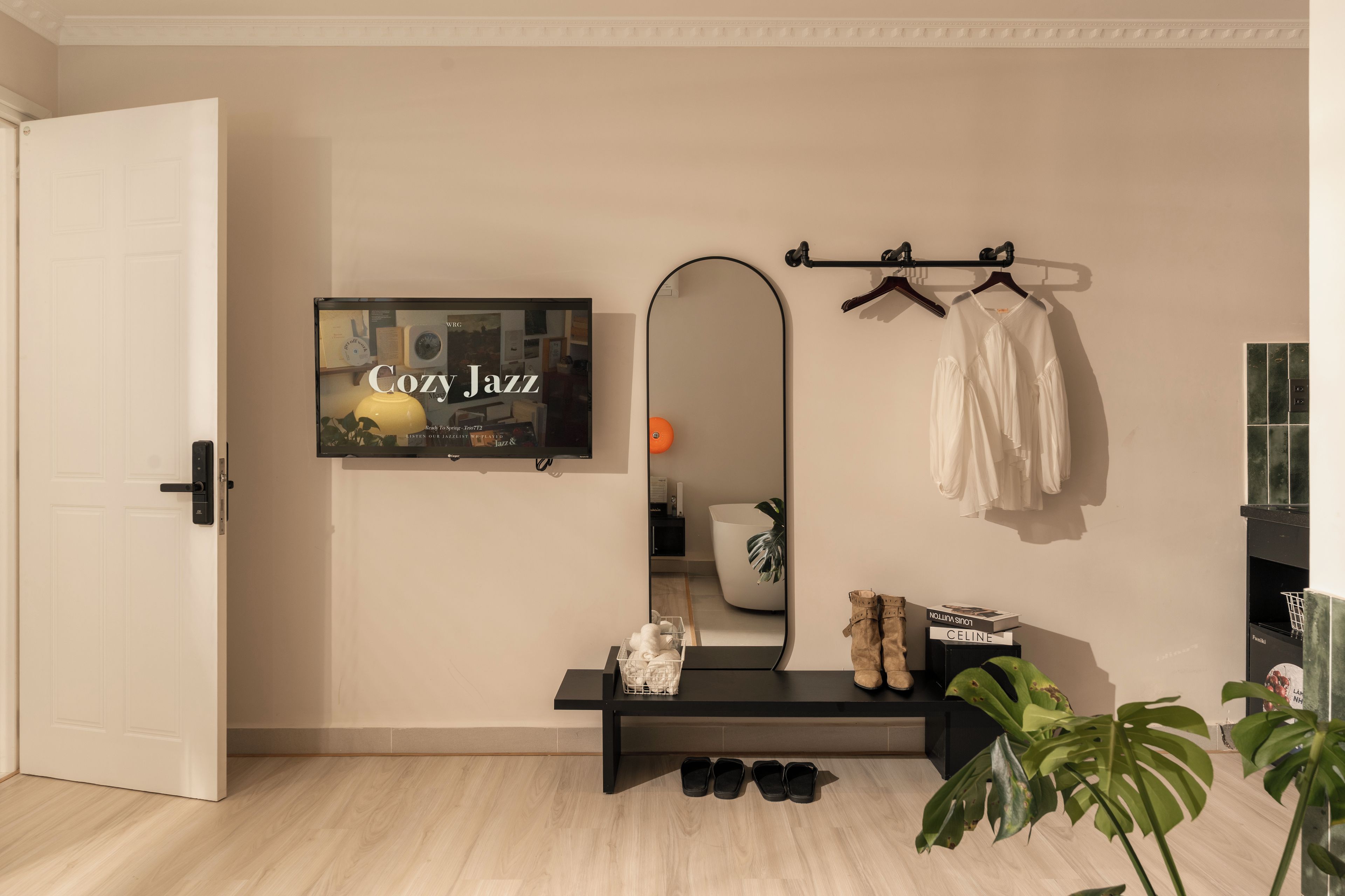Una habitación con una puerta blanca, un banco negro con zapatos, un espejo y un televisor de pared mostrando 'Cozy Jazz'.