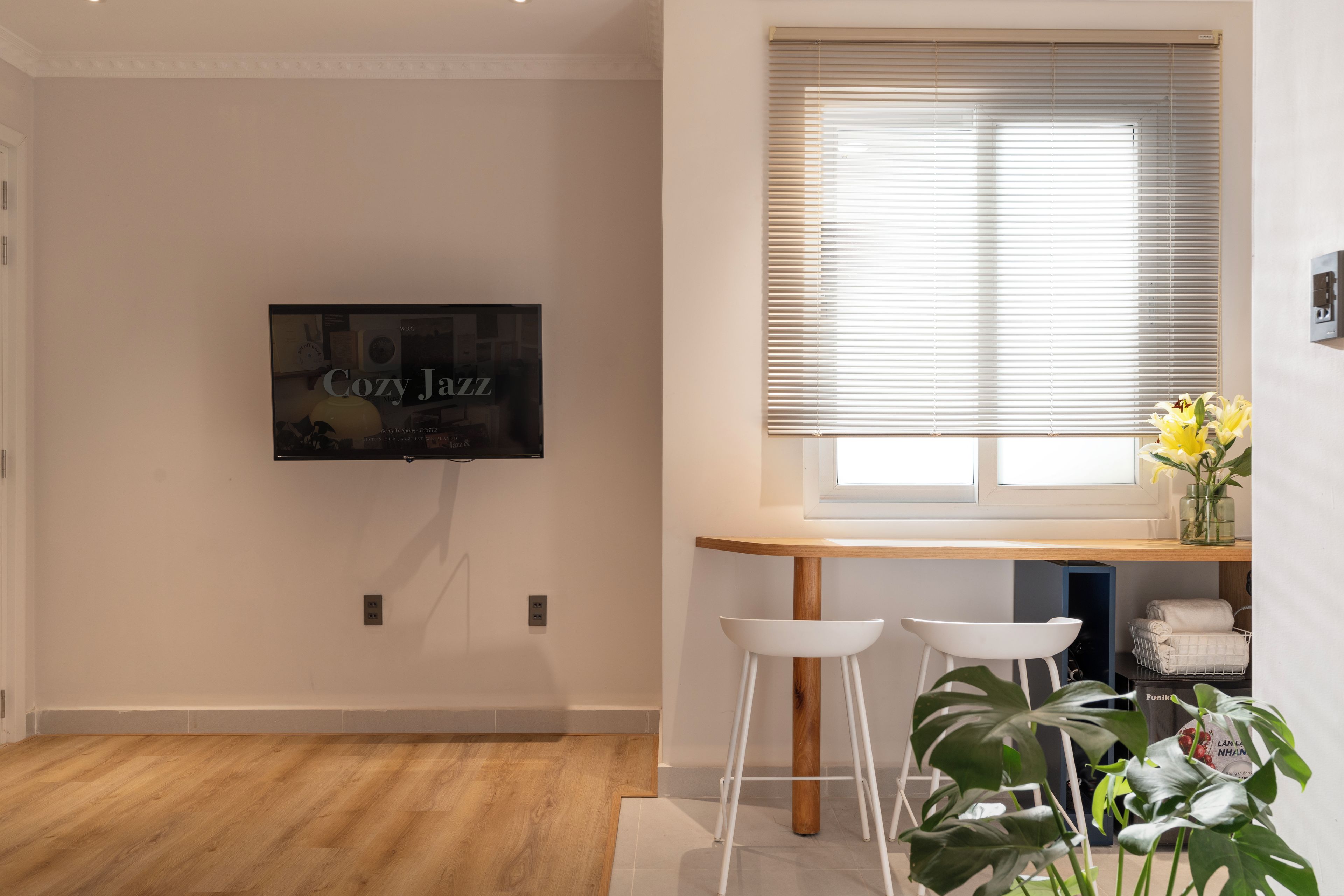 Una sala moderna con un televisor de pantalla plana montado en la pared, una mesa de madera con dos taburetes blancos, una ventana con persianas y una planta en maceta.