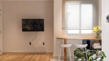 Sala de estar moderna com televisão de tela plana na parede, mesa de madeira com dois banquetes brancos, janela com persianas e planta em vaso.