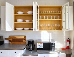 A kitchen with open cabinets containing bowls and glasses, a coffee maker, a microwave, and a fire extinguisher.