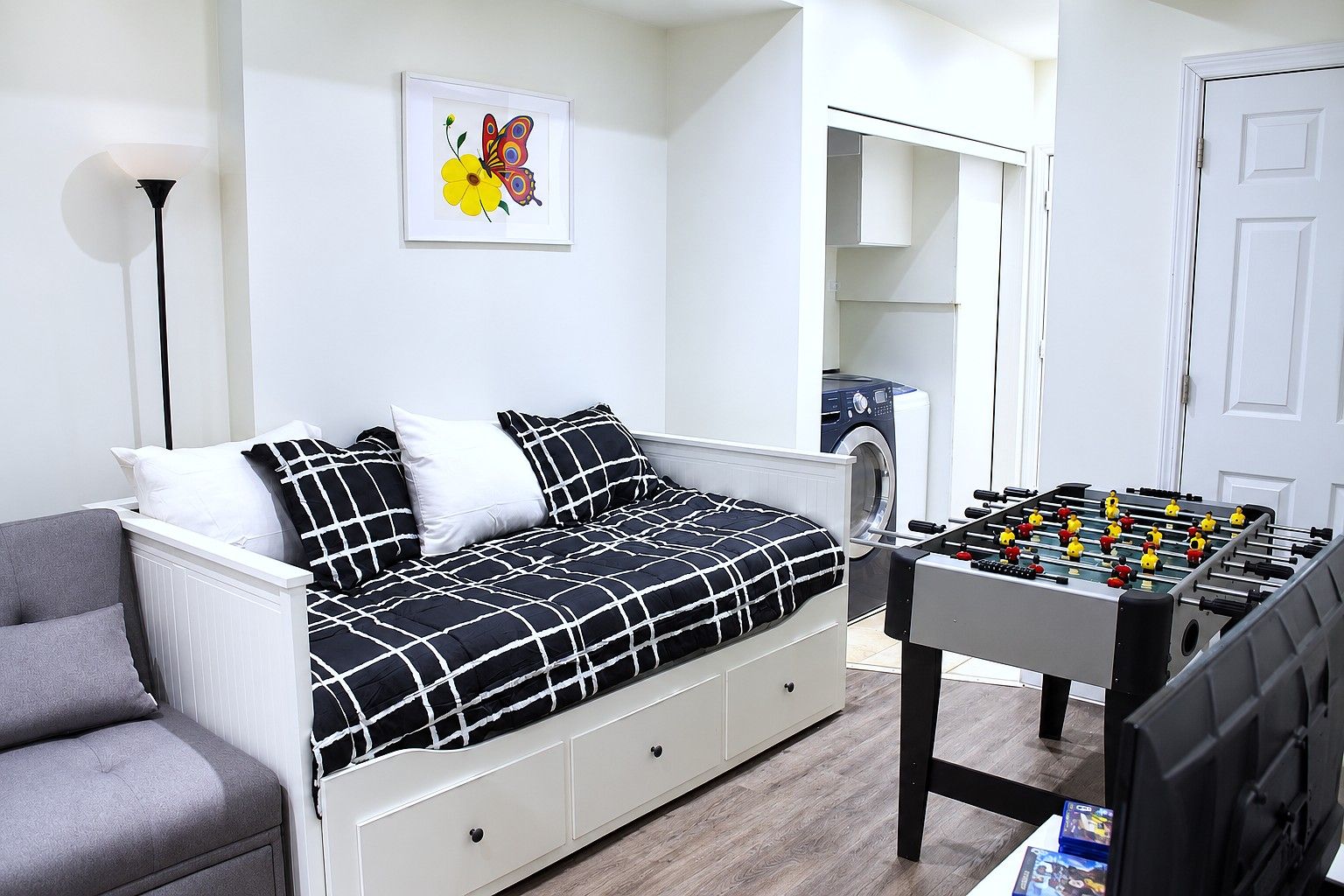 Une chambre avec un lit, un fauteuil gris, une table de baby-foot, un lave-linge et une œuvre d’art représentant un papillon, encadrée et accrochée au mur.