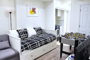 A bedroom with a bed, a grey armchair, a foosball table, a washing machine, and a framed butterfly artwork on the wall.