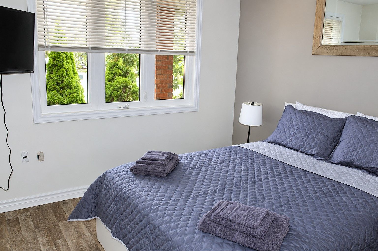 A bedroom with a bed, a TV mounted on the wall, a window with blinds, a bedside lamp, and a mirror on the wall.