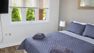 A bedroom with a bed, a TV mounted on the wall, a window with blinds, a bedside lamp, and a mirror on the wall.