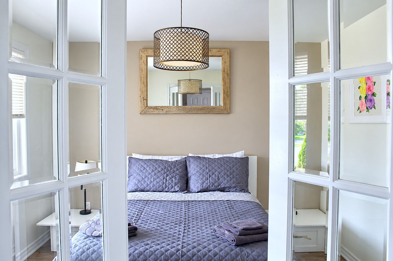 A bedroom with a bed, a hanging light fixture, a mirror, and a window with blinds.
