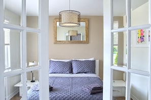 A bedroom with a bed, a hanging light fixture, a mirror, and a window with blinds.