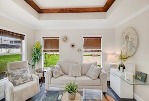Ein Wohnzimmer mit einem beigen Sofa, einem runden Couchtisch und einem Sessel mit Sternenmuster-Kissen. Zwei Fenster mit Jalousien, ein Wanddekor und ein Gemälde sind ebenfalls zu sehen.