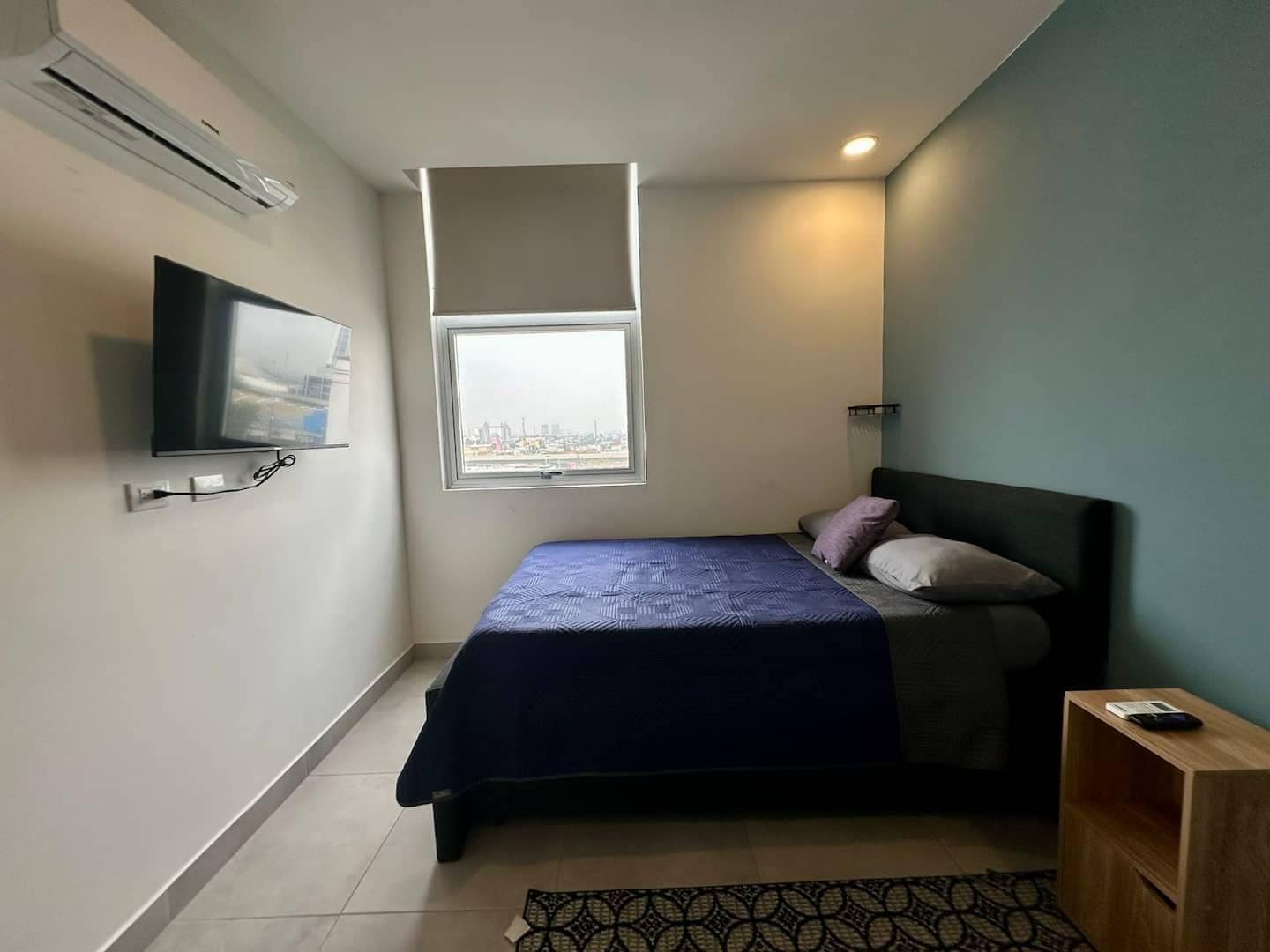 A bedroom with a bed, a television mounted on the wall, a window, and a small wooden side table.