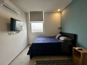 A bedroom with a bed, a television mounted on the wall, a window, and a small wooden side table. - Arena Calzada Apartments (Monterrey)