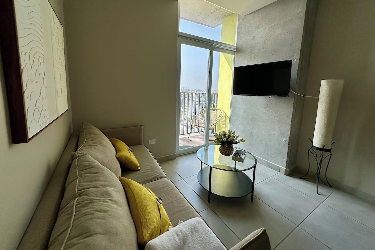 Uma sala de estar moderna com sofá, mesa de centro redonda de vidro, TV na parede e uma varanda com vista.