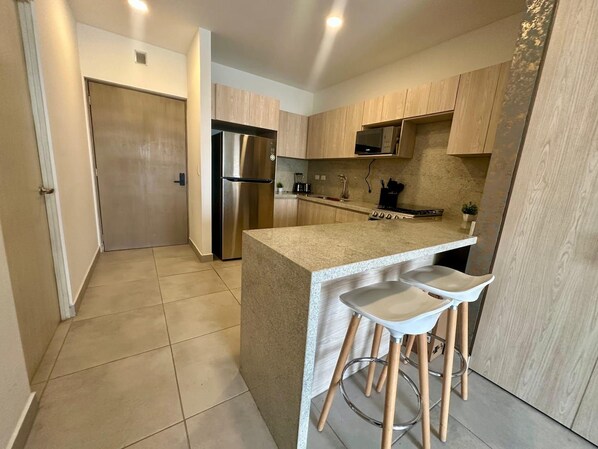 A modern kitchen with a stainless steel refrigerator, wooden cabinets, and a central island with bar stools. - Arena Calzada Apartments (Monterrey)