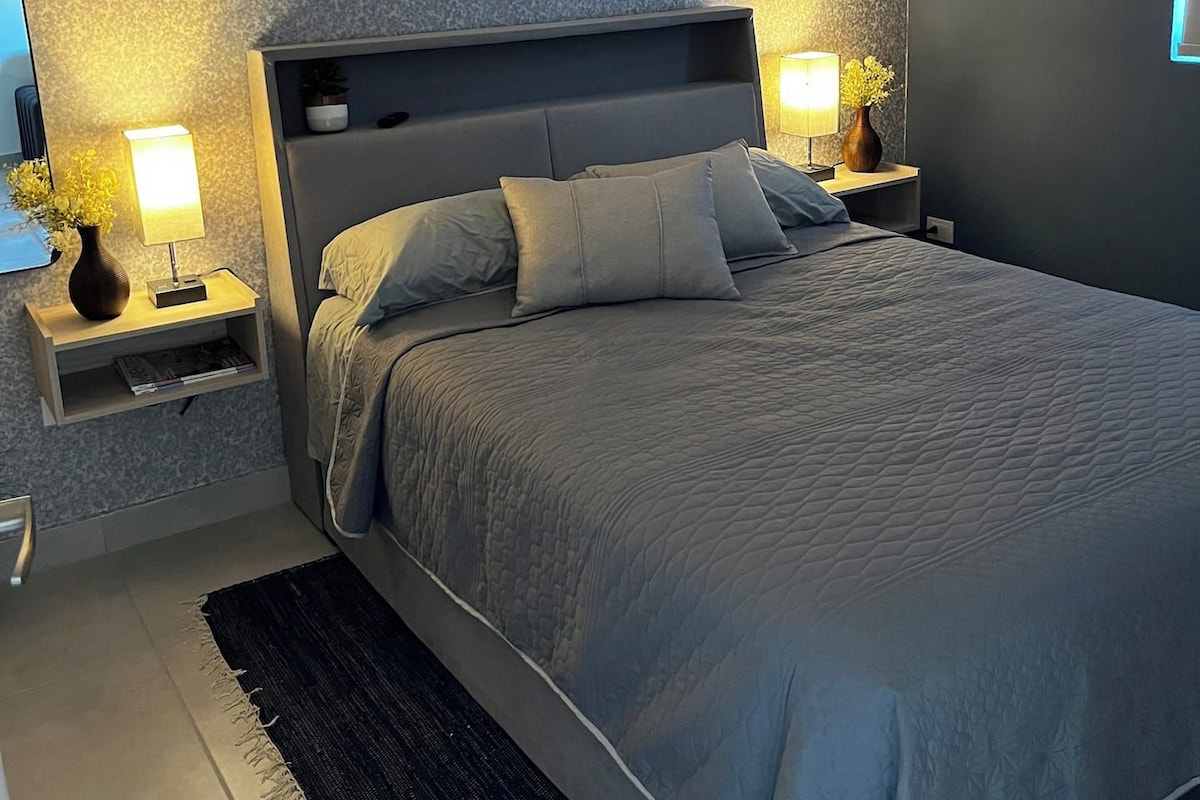 Quarto moderno com cama grande, duas mesas de cabeceira com luminárias, cabeceira escura e parede texturizada.