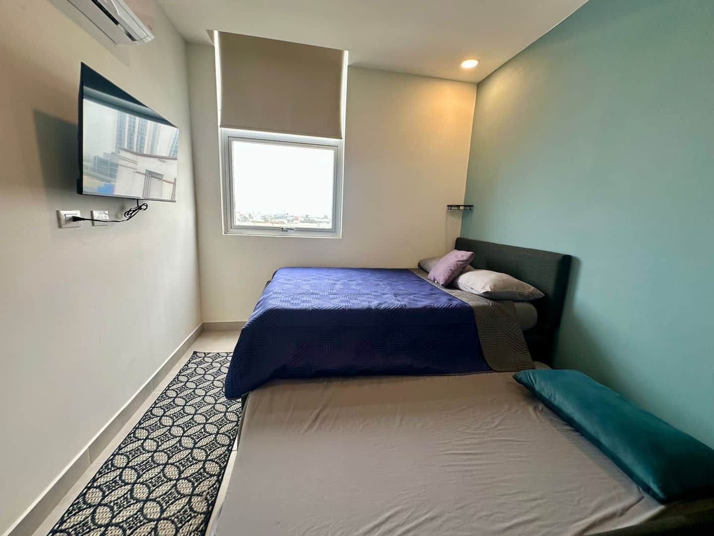 A bedroom with a bed, a window, a wall-mounted TV, and a patterned rug.