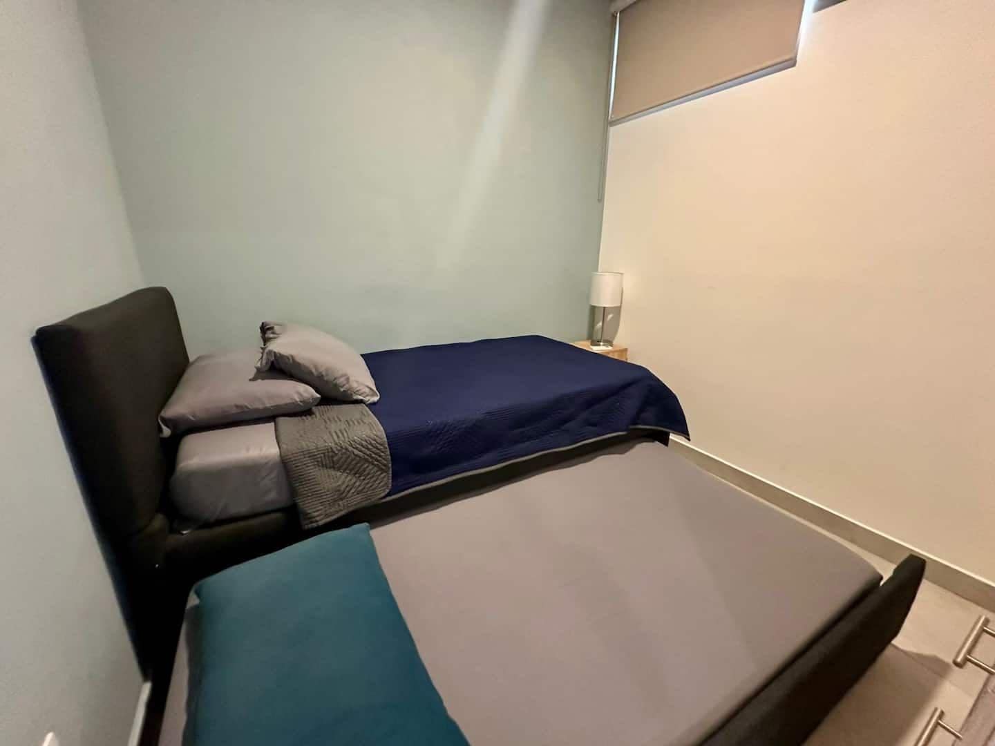 Una cama individual con colchón azul y manta gris, una mesita de noche con lámpara, y un espejo sujeto a la pared.