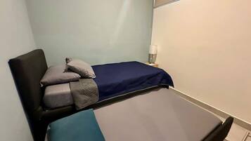 Una cama individual con colchón azul y manta gris, una mesita de noche con lámpara, y un espejo sujeto a la pared.