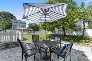 Une terrasse extérieure avec une table et des chaises, un parasol à rayures et une clôture blanche en bois.