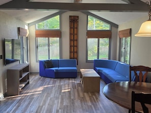 A spacious living room with a large blue sofa, a wooden coffee table, and a TV mounted on the wall. - Caberfae Cabin - 3BR Next to Caberfae Peaks Ski & Golf Resort (Cadillac)