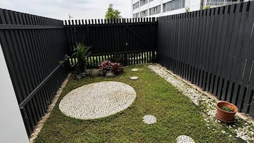 A fenced outdoor area with a circular patterned stone path, a small garden with potted plants, and a building in the background.
