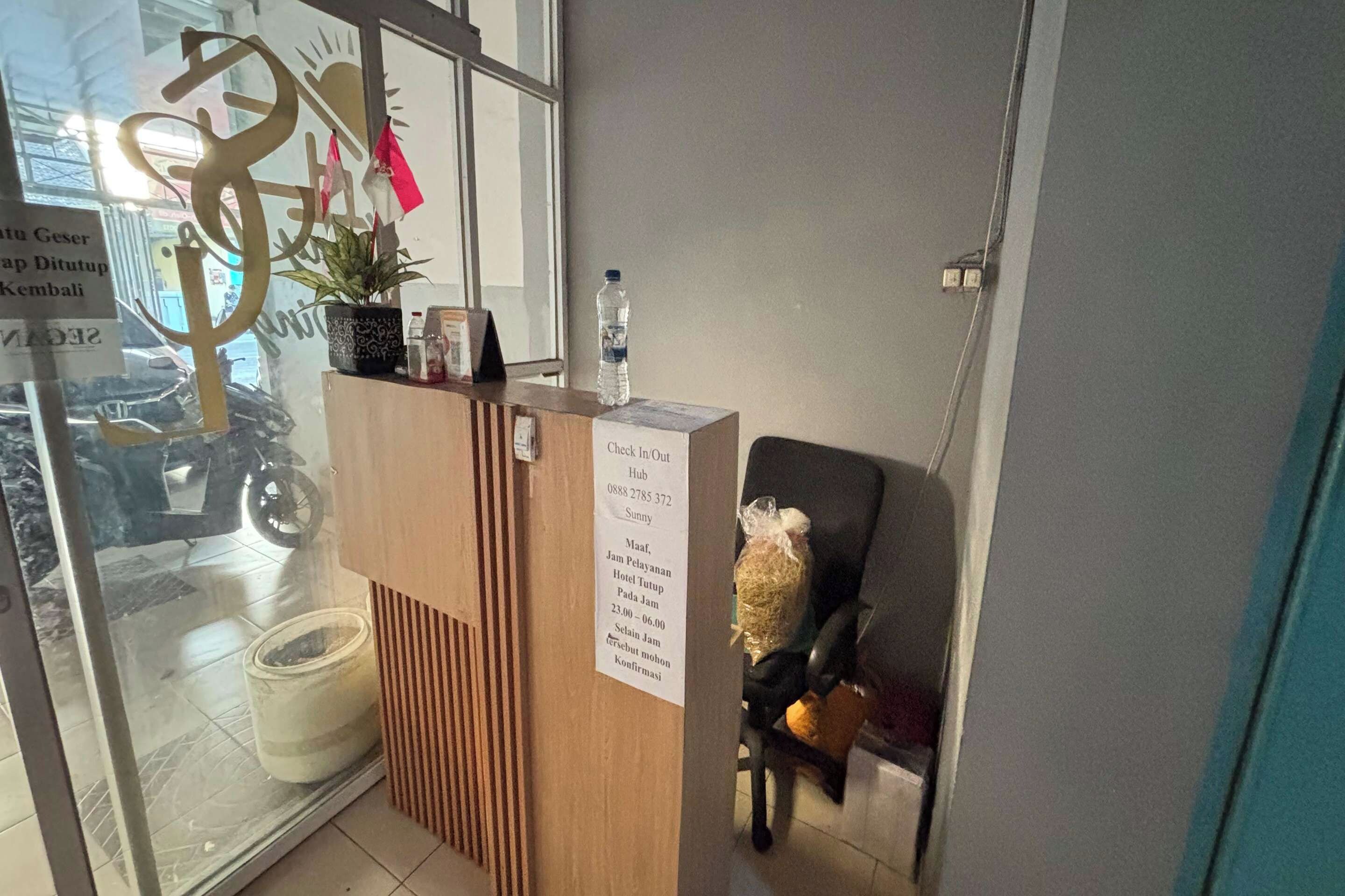 A reception desk with a 'Check In/Out' sign, a water bottle, and a potted plant.