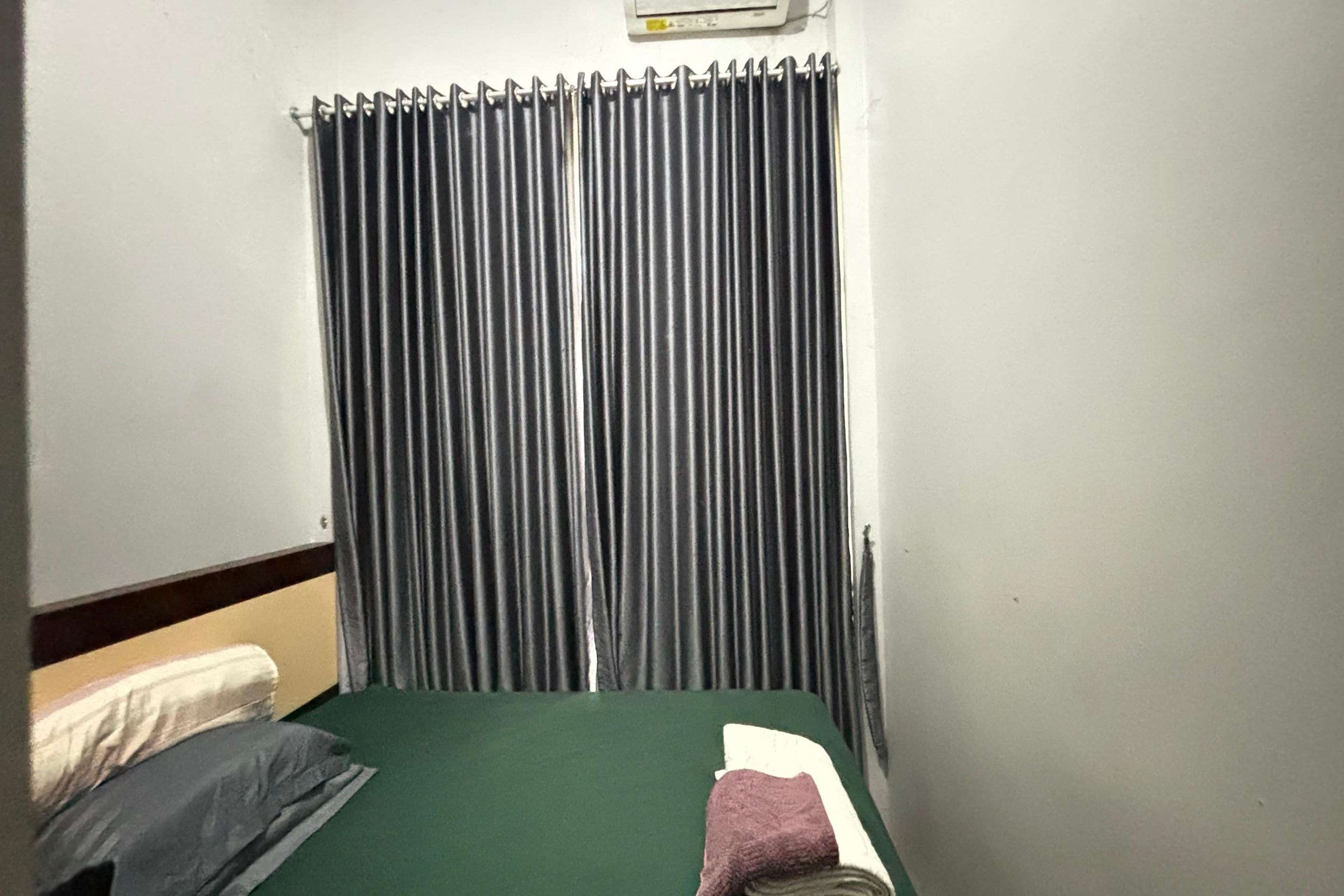A room with a bed, a window with vertical blinds, and a wall-mounted air conditioning unit.