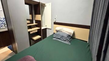 Un letto singolo con materasso verde, un cuscino e una coperta. È presente un comodino con una lampada. Si nota un armadio con specchio e un cappotto appeso.