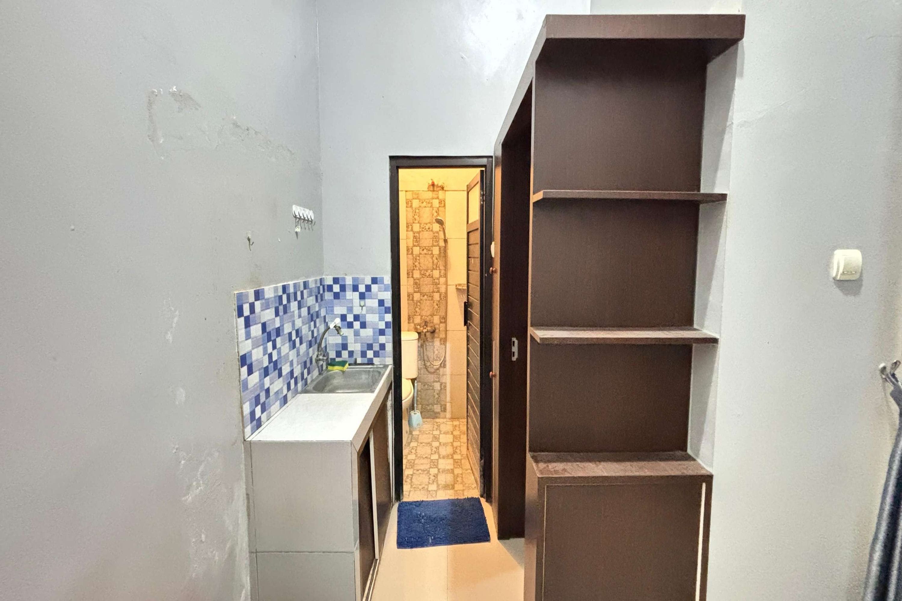 Een smalle gang met aan de linkerkant een keukenhoek, voorzien van een betegelde spatwand en een spoelbak. Aan het einde van de gang is een badkamer met douche en toilet zichtbaar door een geopende deur.