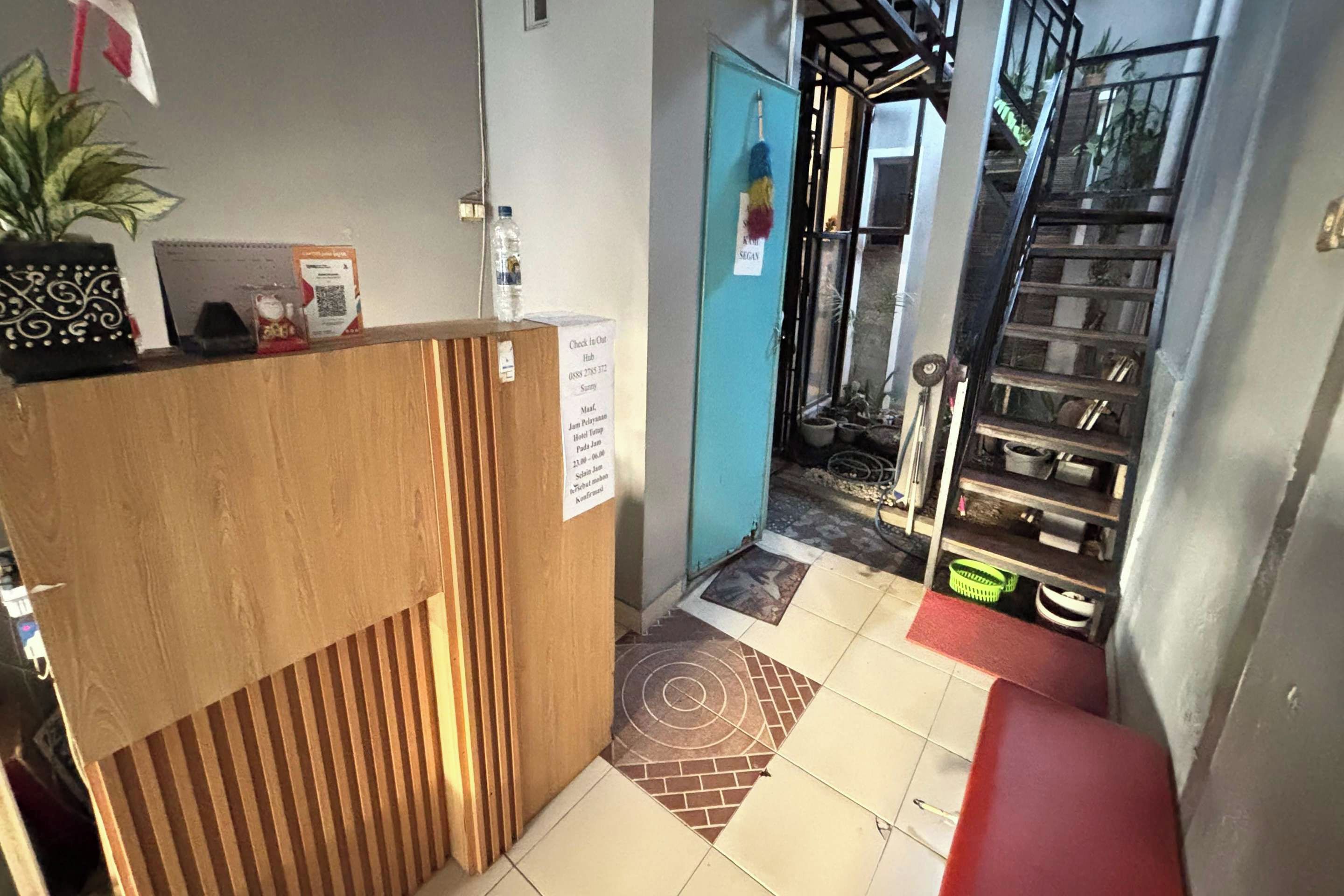 Uma área de recepção de hotel com uma mesa de madeira, uma porta azul, uma escadaria e piso ladrilhado.