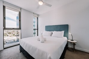 A hotel room with a large bed, a ceiling fan, a bedside table with a lamp, and a view of the city through a large window. - Refined One Bedroom Retreat (Fortitude Valley)