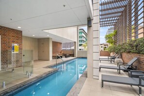 A modern indoor swimming pool with clear glass walls, a seating area with a bench and chairs, and a view of the cityscape through large windows. - Refined One Bedroom Retreat (Fortitude Valley)