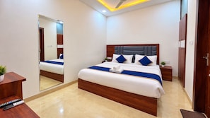 A modern hotel room with a large bed, a wooden headboard, a bedside table with a lamp, and a mirror on the wall. - Hotel Ganga  Palace - Ellara Hotels  (Agra)