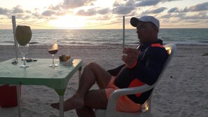 Eine Person sitzt an einem Tisch mit einem Getränk und einem Glas, im Hintergrund ein Strand und das Meer.