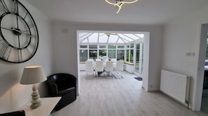 Ein modernes Interieur mit einer großen Uhr, einem Essbereich mit weißer Möbel und einem Blick durch eine Glastür in einen Außenbereich.
