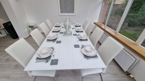 Une salle à manger avec une longue table dressée pour six personnes, des chaises blanches et une grande fenêtre donnant sur un paysage verdoyant.