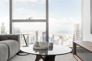 A modern living room with a large window, a round table with a vase of flowers, and a grey sofa. - Sky High 2BR with Spectacular City Views (Melbourne)