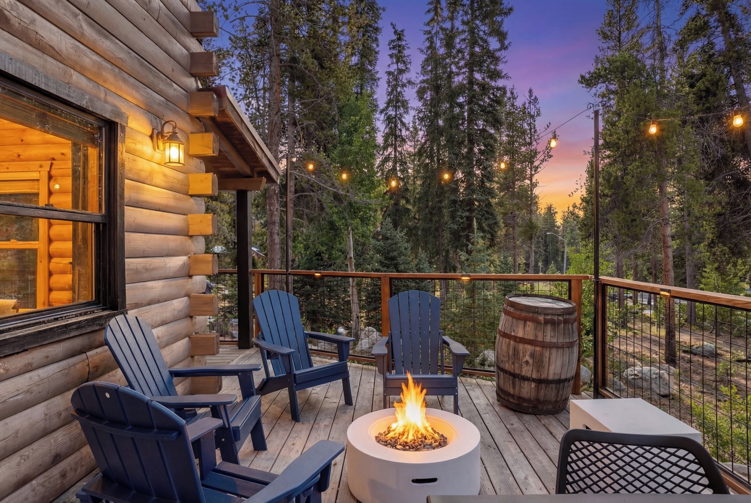 A log cabin with a deck, outdoor seating, a fire pit, and string lights.
