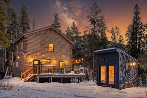 A log cabin with a deck and a small attached structure, surrounded by snow and trees. - 8 min to Breck DT•Steps to Stream•Hottub/Sauna (Breckenridge)