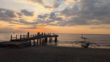 Une jetée s’avançant dans une mer calme, avec des personnes rassemblées à son extrémité, un bateau sur la plage et un coucher de soleil en arrière-plan.