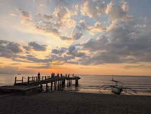 Un muelle se extiende sobre un mar tranquilo, con gente reunida al final, un bote en la orilla y una puesta de sol al fondo.